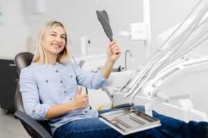 Happy dental patient holding mirror and making thumbs up gesture 
